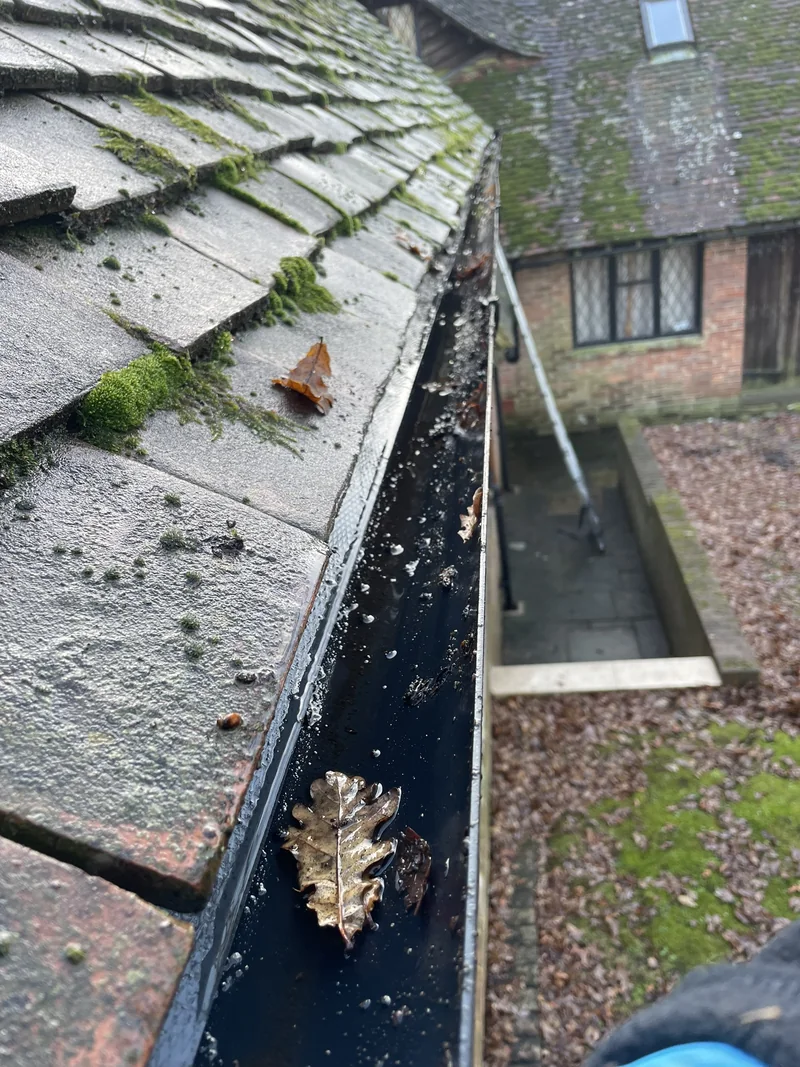 Leaves and residue in a black gutter below mossy roof tiles
