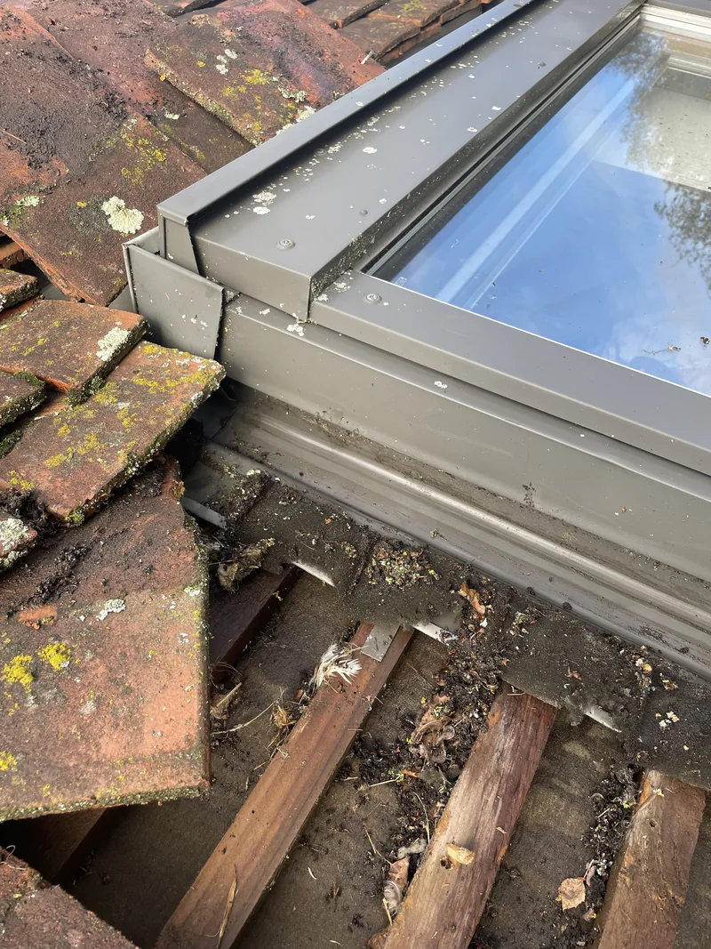 Lifted tiles beside a roof window revealing debris around the battens and flashing