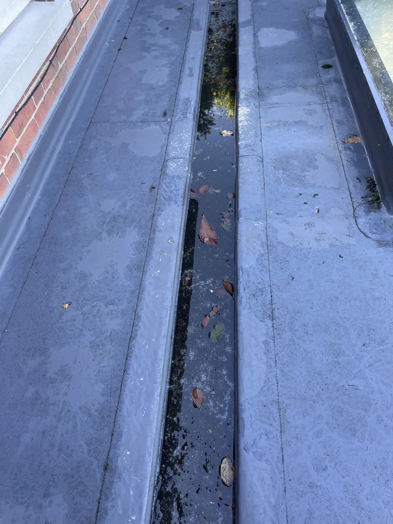 Standing water and scattered debris in a lead-lined box gutter beside brickwork